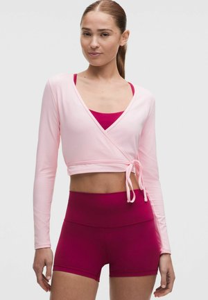 Mujer con camiseta cruzada de manga larga en rosa claro sobre un sujetador deportivo rosa oscuro y pantalones cortos deportivos de cintura alta rosa oscuro a juego.