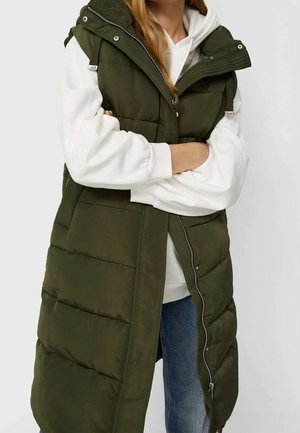Personne portant un long gilet matelassé vert olive par-dessus un sweat à capuche blanc et un jean bleu, les bras croisés à la taille.