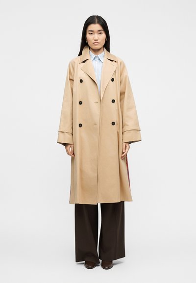 Γυναίκα που φοράει ένα μπεζ διπλόκαμπτο παλτό trench πάνω από μια ριγέ μπλούζα και φαρδιά σκοτεινά παντελόνια, στέκεται μπροστά σε απλό φόντο.