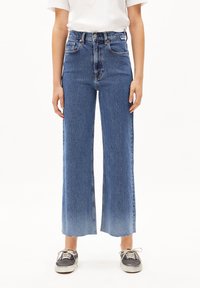 Jeans de mezclilla de tiro alto en lavado azul medio, corte recto, dobladillo sin terminar, con bolsillos delanteros y traseros, combinados con zapatos negros informales.