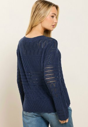 Donna di profilo che indossa un maglione a maglia aperta blu navy con motivi geometrici e jeans azzurri su uno sfondo semplice.