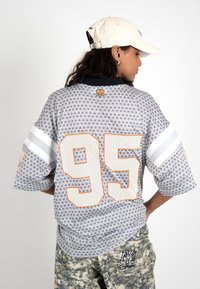 Mujer con camiseta deportiva gris con estampado y número 95 grande con borde naranja, sombrero blanco y pantalones de camuflaje con logo bordado.