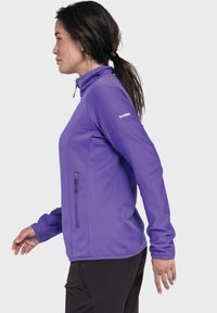 Lila Zip-Up-Jacke mit hohem Kragen, strukturiertem Stoff und einer Reißverschlusstasche auf der Vorderseite. Lange Ärmel mit Bündchen und Markenlogo auf der Schulter.
