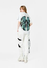 Camisa blanca de gran tamaño con gráficos de alas en azul y negro en la parte posterior, combinada con pantalones jogger blancos que presentan diseños de plumas negras.