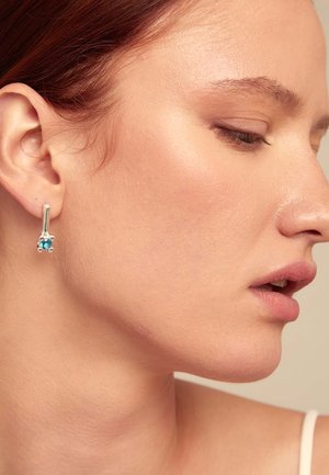 Pendientes de plata con una piedra preciosa turquesa, presentando un diseño cónico, mostrados en la oreja de un modelo en perfil contra un fondo neutro.