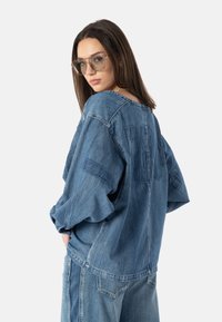 Overdimensjonert denimskjorte i lys blå, med bred halslinning, senkede skuldre og "CMM" brodert på ryggen. Teksturert stoff.