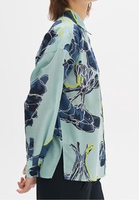 Long-sleeve shirt in light mint green fabric with a floral pattern in navy blue and yellow accents, featuring a classic collar and side slits.