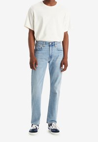 Personne portant un t-shirt blanc uni à manches courtes, un jean bleu clair droit et des baskets blanches et bleu marine, debout devant un fond blanc.