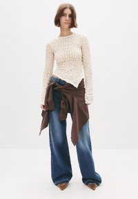 Vrouw met kort haar in een crèmekleurige, gestructureerde longsleeve top, wijde blauwe jeans en een bruine jas om haar taille geknoopt.
