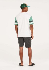 T-shirt blanche avec des manches vertes à rayures, associé à un short gris et des sandales noires, vu de dos, avec une casquette verte.