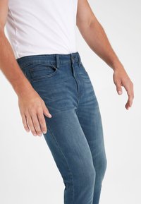 Eng anliegende blaue Jeans aus Denim mit leichtem Stretch, fünf Taschen-Design, Reißverschluss und dezenten Abnutzungserscheinungen in der Nähe der Oberschenkel.