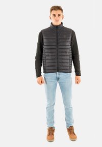 Zwarte gewatteerde bodywarmer met horizontale stiknaden, hoge kraag en volledige ritssluiting, gedragen over een zwart shirt met lange mouwen en lichtblauwe jeans.