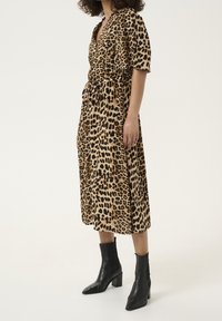 Femme portant une robe midi à imprimé léopard avec manches courtes, taille nouée, et bottines noires à talons sur un fond blanc uni.