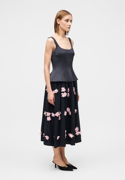 Τοπ πεπλούμ από σκουρόχρωμο γκρι ύφασμα με εφαρμοστό μπούστο· ολόκληρη μαύρη φούστα διακοσμημένη με ροζ λουλουδάτες εφαρμογές. Μαύρα παπούτσια με μυτερή μύτη.