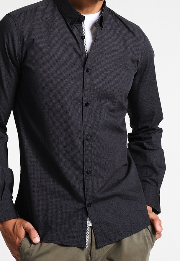 Homme portant une chemise texturée gris foncé à boutons sur un sous-vêtement blanc avec un pantalon kaki, les mains dans les poches, sur un fond uni.