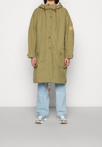 Długa parka w kolorze oliwkowej zieleni z kapturem ze sznurkiem, zatrzaskami i dwoma przednimi kieszeniami. Nosi się ją na jasnoniebieskich szerokich dżinsach i trampkach.