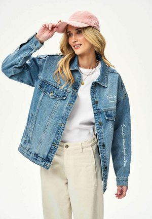 Blondínka v svetlom modrom rifľovom sakom s bielym textom, bielom tričku, béžových nohaviciach a ružovej čiapke, držiaca šilt čiapky jednou rukou.