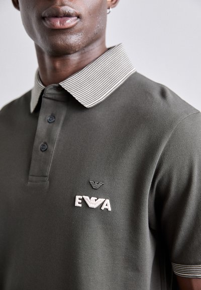 Olīvu zaļš polo krekls ar strīpotu apkakli, trim pogām un izšūtu logotipu krūtīs baltā un melnā krāsā. Gluda auduma tekstūra.