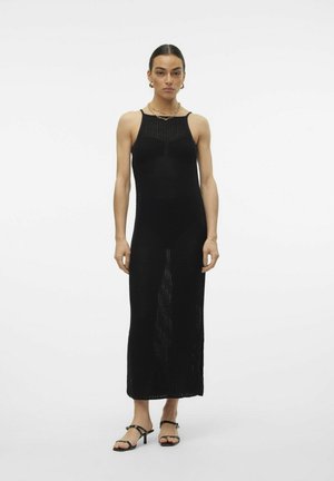 Abito maxi nero a maglia con spalline sottili, caratterizzato da un motivo texturato e uno spacco laterale. Abbinato a sandali con tacco a strappo neri.