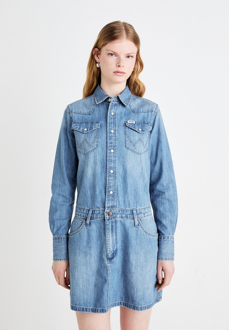 Ανοιχτού μπλε τζιν φόρεμα με κουμπιά μπροστά, με μυτερό γιακά, δύο τσέπες στο στήθος και καθορισμένη μέση. Διαθέτει μακρύ μανίκι και κυρτό στρίφωμα.