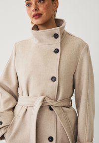 Beige wollen blendjas met een hoge kraag, zwarte knopen, een strikceintuur en zijzakken. Zachte textuur en een casual pasvorm.