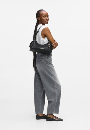 Kobieta z długimi warkoczami, ubrana w białą podkoszulkę bez rękawów, luźne szare dżinsy, czarne płaskie buty, trzymająca czarną torbę na ramię na białym tle.