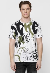 Camiseta de manga corta y cuello redondo en blanco, con patrones de estilo grafiti en negro y verde oliva, y texturas de salpicaduras.