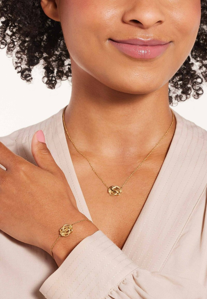 Gold necklace and bracelet with intertwined knot design, worn on a model's neck and wrist. Smooth texture, delicate chain with glossy finish.