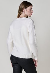 Weißer Strickpullover mit geripptem Saum, versehen mit strukturierten Stickakzenten an den Ärmeln. Das Model kombiniert ihn mit schwarzen Hosen.