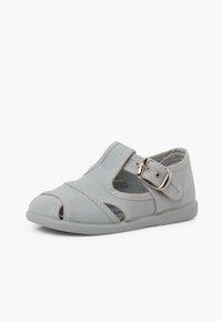 Chaussure en toile grise avec un design ouvert à l'avant, une sangle Velcro et une boucle décorative. La semelle est gris clair avec une structure flexible.