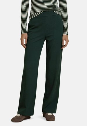 Personne portant un pantalon large vert foncé, des baskets marron et un haut rayé noir et blanc à manches longues, debout devant un fond uni.