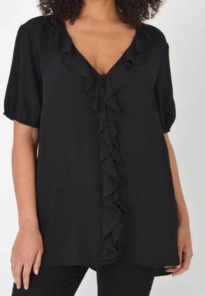 Frau trägt eine schwarze Bluse mit kurzen Ärmeln, V-Ausschnitt und gerafftem Besatz entlang der vorderen Mitte, kombiniert mit schwarzen Hosen.