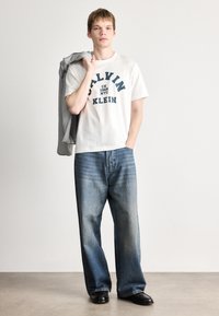 Valge puuvillane T-särk sinise tekstiga, koos helesiniste laia säärega teksade ja mustade kingadega, hoides halli sviitrit õlal.