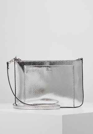 Pochette poignet argentée brillante avec motif serpent texturé, poche frontale rectangulaire et bandoulière amovible, posée sur une surface blanche devant un fond gris.