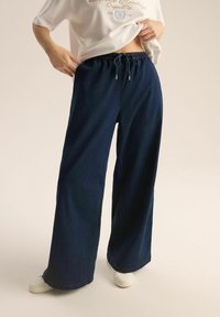 Jeansowe spodnie w szerokim kroju w ciemnoniebieskim kolorze z elastycznym paskiem i sznurkiem. W zestawieniu z białymi sneakersami tworzą swobodny look.