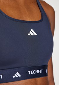 Marineblauwe sport-bh met een ronde halslijn, voorzien van het witte adidas-logo en een "TECHFIT"-band. Smalle stofstructuur. Close-up weergave.