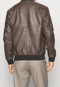 Giacca bomber marrone in pelle con colletto e polsini neri a coste. Texture liscia, con dettagli di cuciture orizzontali sul retro. Adatta per un uso casual.