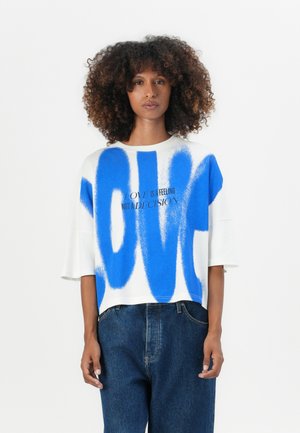 T-shirt bianco corto con testo spray blu "LOVE" e la frase "L'AMORE È UNA SENSAZIONE NON UNA DECISIONE" in nero.