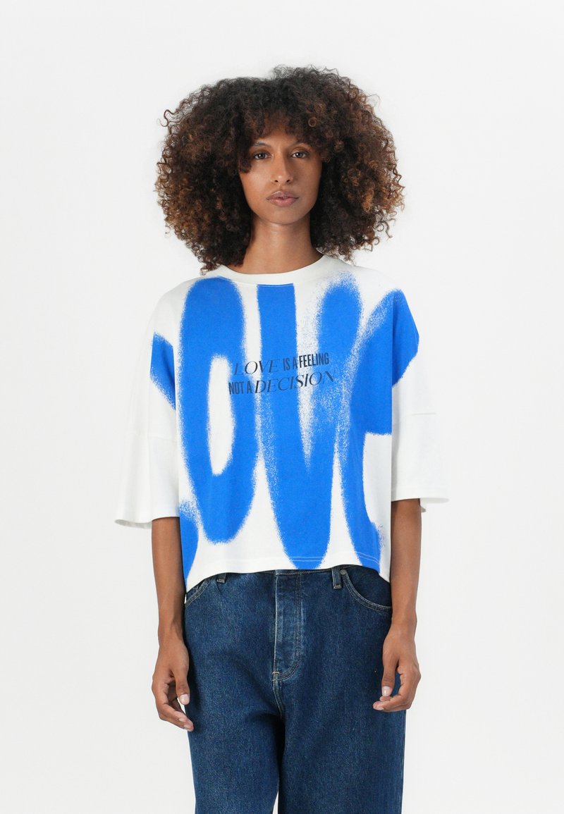 White cropped t-shirt featuring blue spray-painted text "LOVE" with the phrase "LOVE IS A FEELING NOT A DECISION" in black.