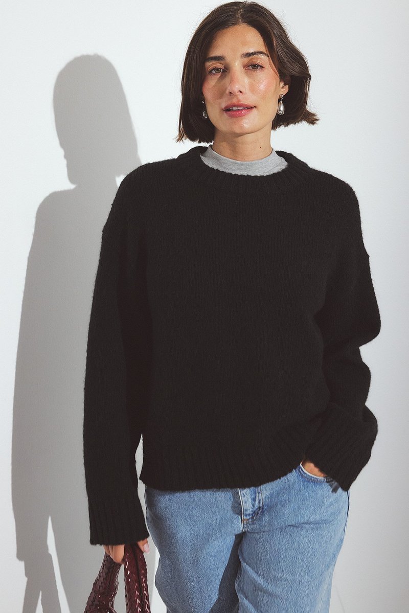 Schwarzer Strickpullover mit geripptem Saum und Bündchen, getragen über einem grauen T-Shirt. Kombiniert mit hellblauen Jeans und einer bordeauxfarbenen geflochtenen Handtasche.