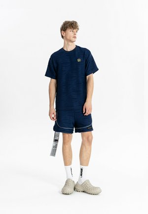 Joven con pantalones cortos texturizados azul marino y camiseta con etiqueta, calcetines blancos y zapatos beige sin cordones, de pie frente a un fondo blanco.