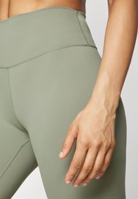 Legginsy o wysokim stanie w oliwkowym kolorze, wykonane z elastycznego materiału, o gładkiej fakturze i minimalistycznych detalach szwów, z ręką opartą na udzie.