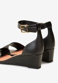 Sandales compensées texturées noires avec brides à la cheville et boucles dorées, bout ouvert et talon fermé, vues de côté et de dos.