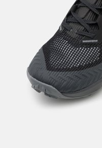 Chaussure de sport noire avec une tige en maille tissée texturée et une semelle en caoutchouc avec un motif en zigzag. Accents gris foncé distincts visibles sur les orteils.