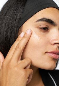 En klar gel appliceras på kinden med fingertoppen. Subjektet har slät, ljusbrun hud och långt svart hår, iklädd ett svart pannband.