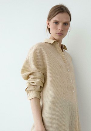 Jeune femme à la peau claire et aux yeux bleus portant une chemise beige à manches longues boutonnée avec les manches retroussées sur un fond blanc uni.