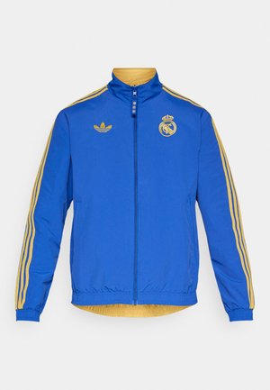 Jachetă albastră cu fermoar, cu accente galbene și logo adidas. Prezintă trei dungi aurii pe fiecare mânecă și un emblema Real Madrid pe piept.