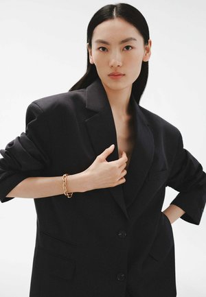 Blazer negro de gran tamaño con hombros estructurados, cierre de un solo botón y dos bolsillos frontales. La modelo lleva una pulsera de cadena dorada.