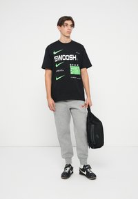 Melna kokvilnas T-krekls ar uzrakstu drosmīgā zaļā un baltā krāsā, kā arī Nike logotipiem, apvienots ar pelēkiem sporta biksēm, kas rotātas ar oranžu logotipu. Rokā melna soma.