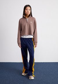 Sweatshirt castanho com fecho éclair e gola alta, combinado com calças de treino azul-marinho com riscas amarelas dos lados. O modelo usa ténis claros.